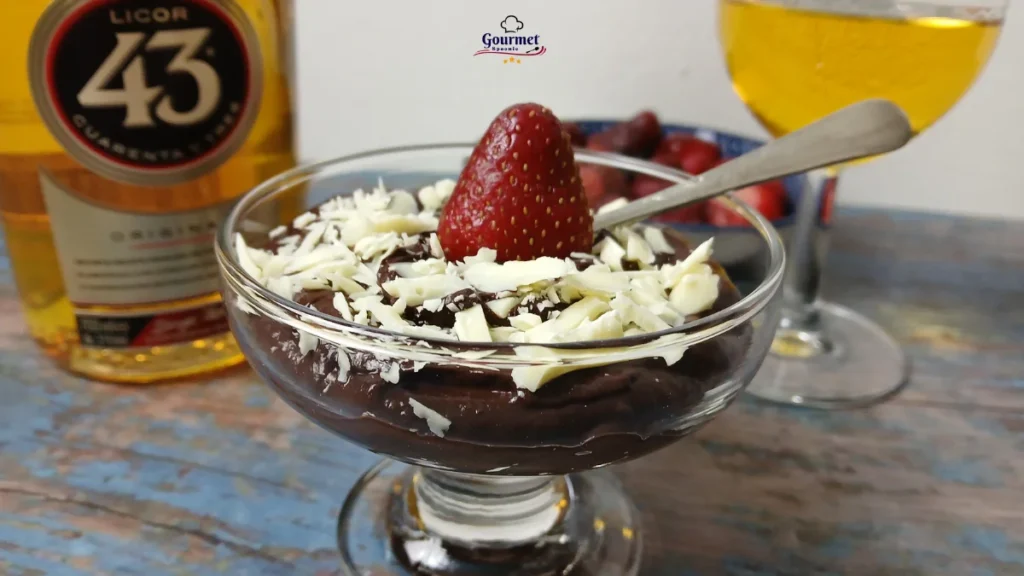A glass dessert dish with chocolate mousse, topped with white chocolate shavings and a strawberry, sits on a wooden table next to a bottle of Licor 43 and a glass of golden beverage. This is an indulgent dessert experience.