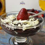 A glass dessert dish with chocolate mousse, topped with white chocolate shavings and a strawberry, sits on a wooden table next to a bottle of Licor 43 and a glass of golden beverage. This is an indulgent dessert experience.