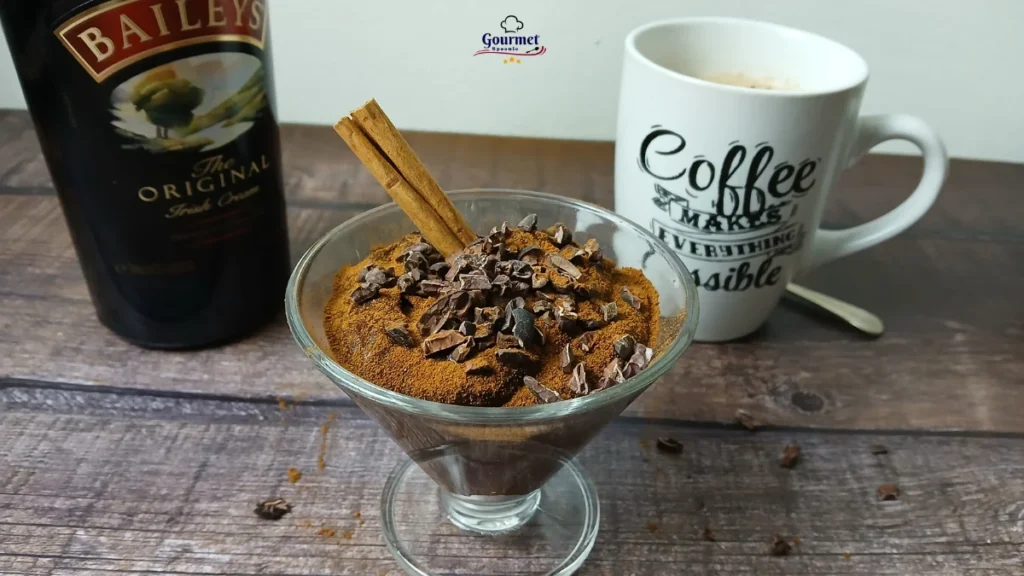 A glass of chocolate mousse topped with cocoa powder, chocolate shavings, and a cinnamon stick sits on a wooden surface. A bottle of Baileys and a mug labeled “Coffee makes everything possible” are beside it.