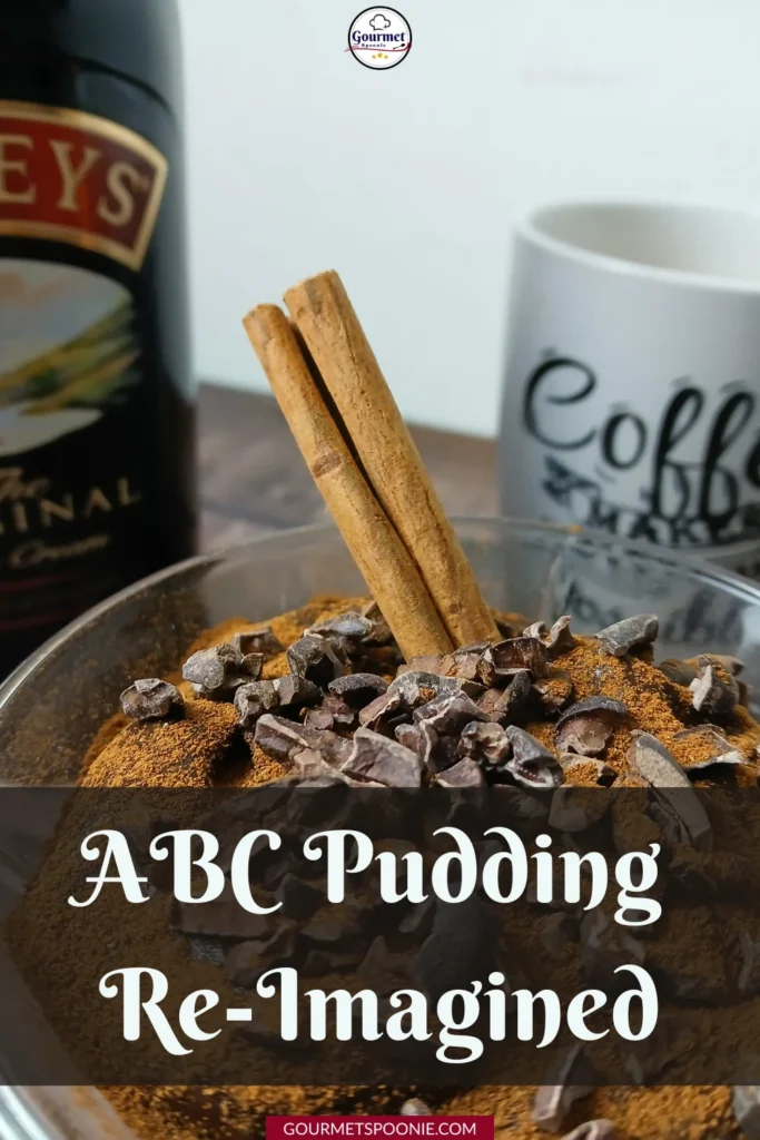 A close-up of a dessert topped with cocoa nibs, cinnamon powder, and two cinnamon sticks, with a bottle of Baileys and a coffee mug in the background. Text overlay reads ABC Pudding Re-Imagined.