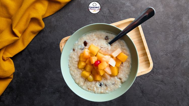 A bowl of oatmeal topped with assorted diced fruits, including mango and a cherry, sits on a wooden tray with a spoon. Perfect for quick healthy meals, its set beside a yellow cloth on a dark stone surface.