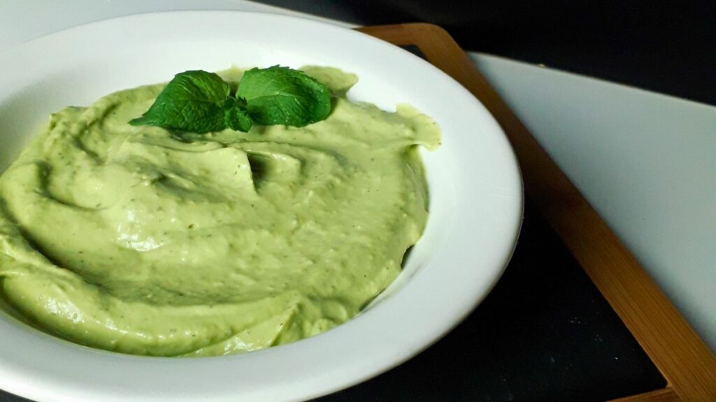 A white bowl filled with creamy green Easy Vegan Avocado Mayonnaise, garnished with two fresh mint leaves, sits on a dark wooden tray against a light-coloured background.