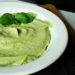 A white bowl filled with creamy green Easy Vegan Avocado Mayonnaise, garnished with two fresh mint leaves, sits on a dark wooden tray against a light-coloured background.