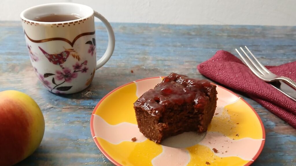 A cup of tea, a floral mug, and gluten-free chickpea brownies with jam on a colorful plate sit on a blue wooden table. An apple, red napkin, and silverware are nearby.