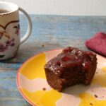 A cup of tea, a floral mug, and gluten-free chickpea brownies with jam on a colorful plate sit on a blue wooden table. An apple, red napkin, and silverware are nearby.