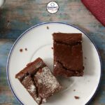 A plate with two pieces of vegan cherry chocolate brownies—one dusted with sugar—sits on a rustic blue table. Nearby are a red apple, a cup of tea, a red napkin, and a fork and knife.