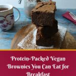 A stack of chocolate brownies dusted with cocoa powder sits on parchment paper, with a floral teacup, a glass jar of brownies, and red napkins on a rustic blue wooden table. Text reads: Easy Vegan Baking—Protein-Packed Brownies You Can Eat for Breakfast.