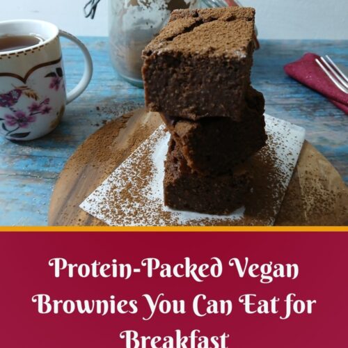 A stack of chocolate brownies dusted with cocoa powder sits on parchment paper, with a floral teacup, a glass jar of brownies, and red napkins on a rustic blue wooden table. Text reads: Easy Vegan Baking—Protein-Packed Brownies You Can Eat for Breakfast.