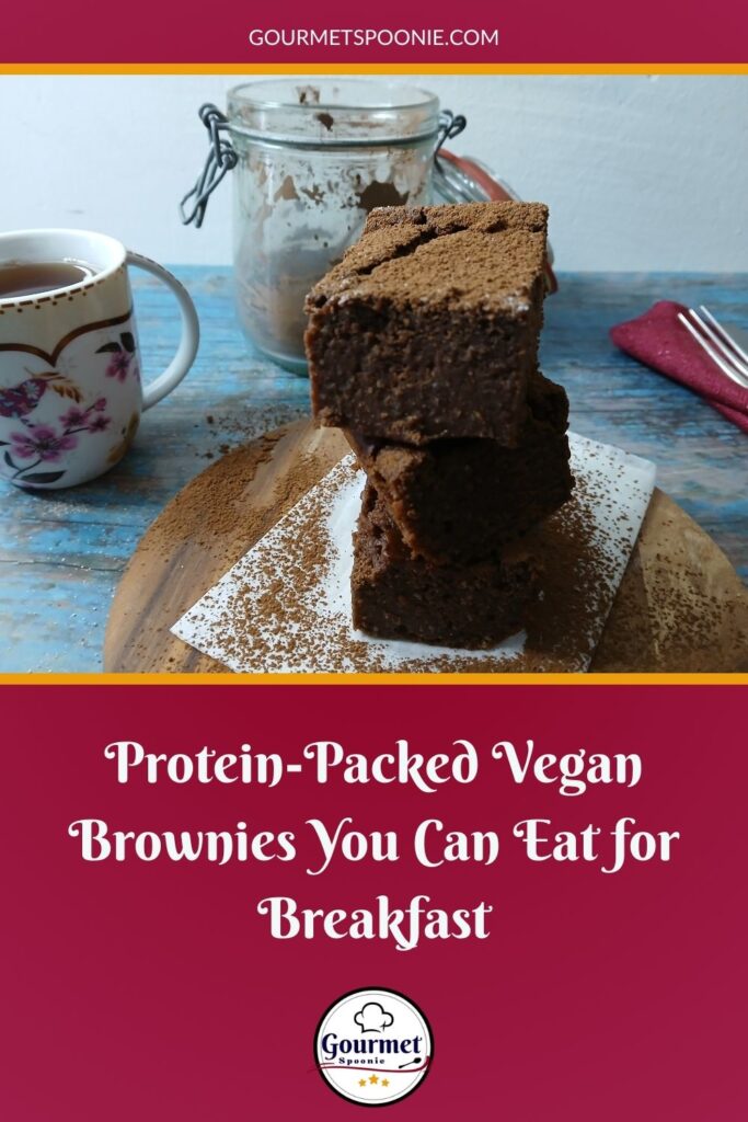 A stack of chocolate brownies dusted with cocoa powder sits on parchment paper, with a floral teacup, a glass jar of brownies, and red napkins on a rustic blue wooden table. Text reads: Easy Vegan Baking—Protein-Packed Brownies You Can Eat for Breakfast.