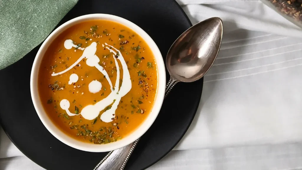 A bowl of vegan carrot and ginger soup, garnished with cream and herbs, sits on a black plate with a silver spoon, next to a green serviette.