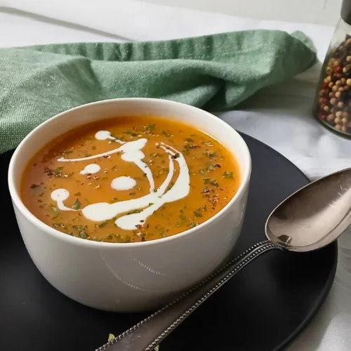A bowl of instant pot carrot and ginger soup with a swirl of cream and herbs on top, placed on a black plate with a spoon beside it; a green serviette and pepper grinder in the background.