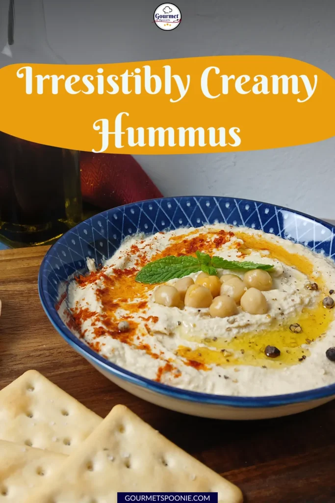 A bowl of creamy hummus topped with whole chickpeas, olive oil, paprika, herbs, and a mint leaf, served with crackers on a wooden surface. Text above reads Irresistibly Creamy Hummus.