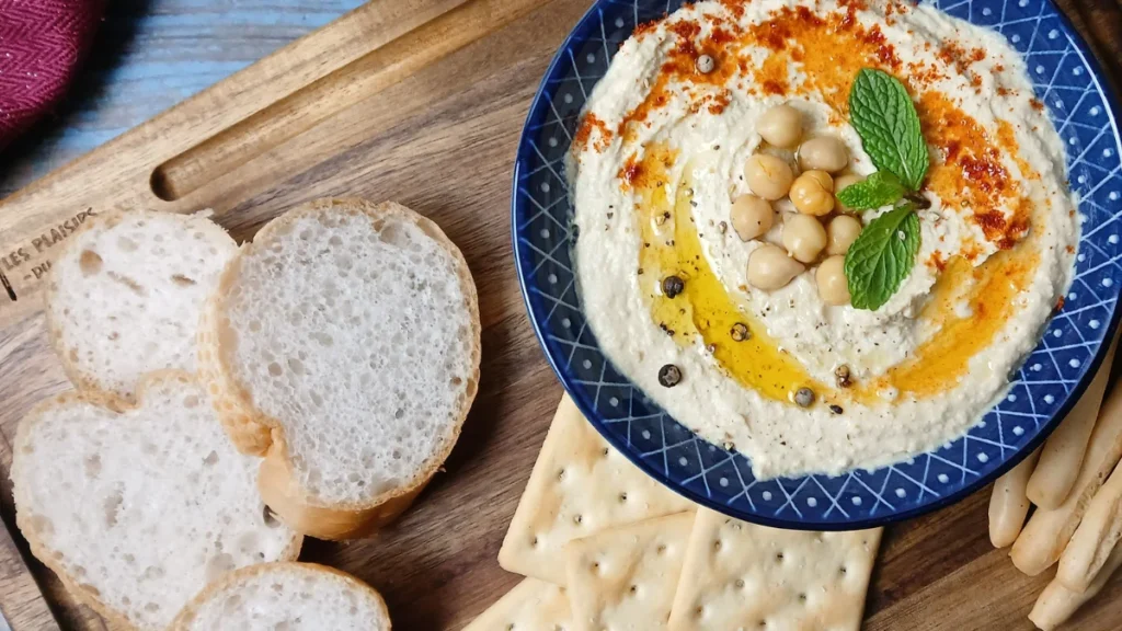 A blue bowl of creamy hummus garnished with chickpeas, mint, olive oil, and spices sits on a wooden board alongside slices of baguette, crackers, and breadsticks.