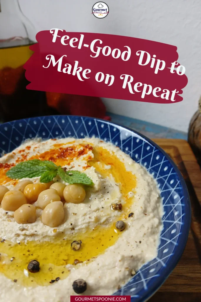 A blue patterned bowl of hummus, garnished with whole chickpeas, olive oil, herbs, and spices, sits on a wooden surface. Text above reads, Feel-Good Dip to Make on Repeat.