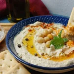 A bowl of creamy hummus garnished with chickpeas, olive oil, spices, and herbs sits on a wooden tray with crackers and sliced bread; a hand dips a cracker into the hummus.