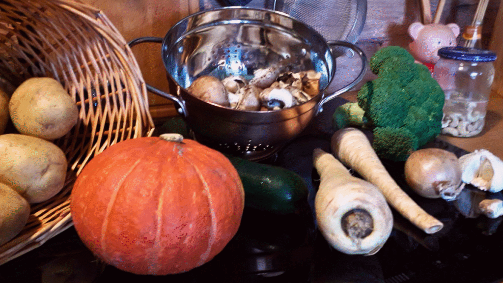A variety of vegetables for meal prepping sit on a kitchen worktop, including potatoes, a pumpkin, cucumber, parsnips, broccoli, mushrooms in a metal colander, and garlic, with a woven basket and jar in the background.