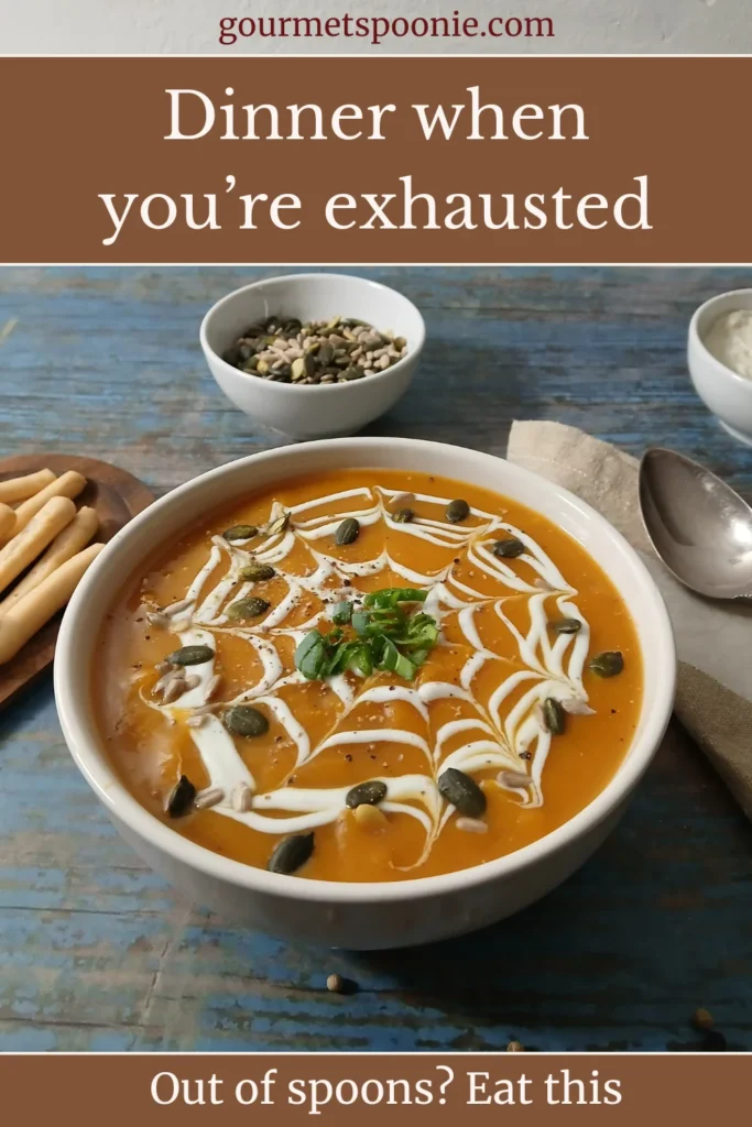 A bowl of orange soup topped with cream in a web pattern, seeds, and herbs sits on a table next to breadsticks, a bowl of seeds, and a spoon.