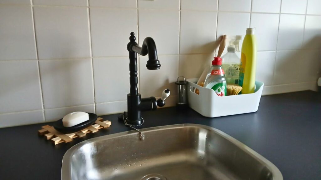 A kitchen sink with a black faucet, a soap dish with white soap, and a white container holding dish soap, a sponge, and cleaning supplies sits on a dark countertop—showcasing easy kitchen cleaning tips for Spoonies against a white tiled backsplash.