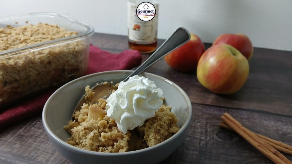 A bowl of easy gluten-free apple crumble topped with whipped cream sits next to a baking dish of crumble, fresh apples, cinnamon sticks, and a bottle in the background on a wooden surface.