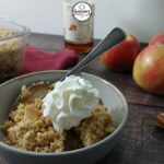 A bowl of easy gluten-free apple crumble topped with whipped cream sits next to a baking dish of crumble, fresh apples, cinnamon sticks, and a bottle in the background on a wooden surface.