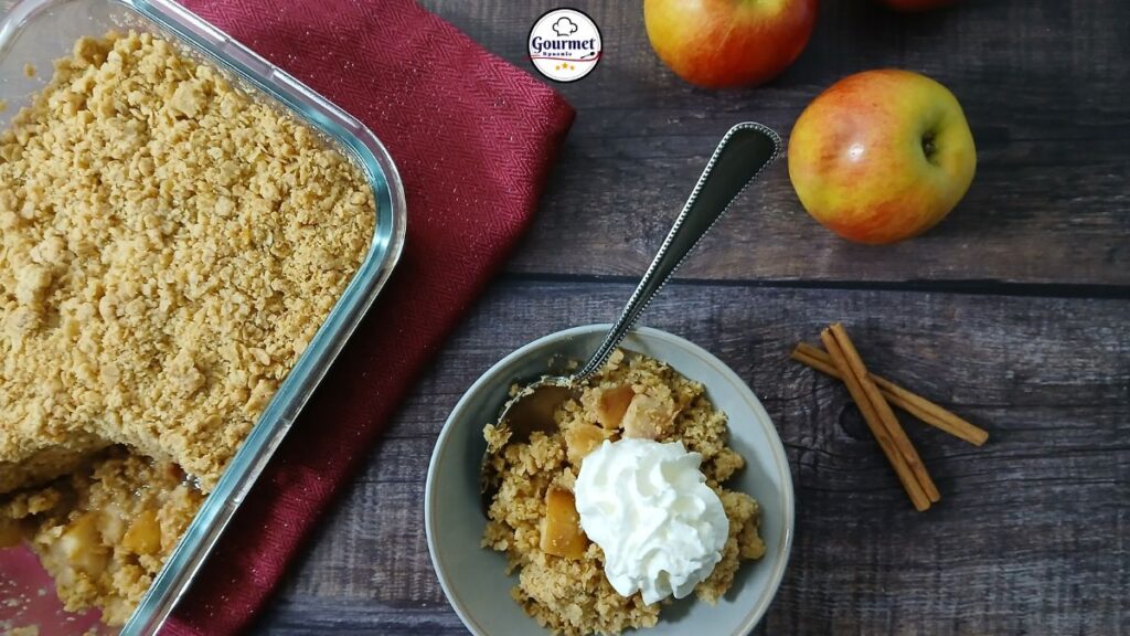 A bowl of easy gluten-free apple crumble topped with whipped cream sits beside a baking dish of crumble, fresh apples, cinnamon sticks, and a red napkin on a wooden table. A spoon rests in the bowl.