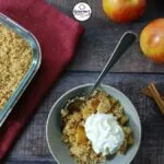 A bowl of easy gluten-free apple crumble topped with whipped cream sits beside a baking dish of crumble, fresh apples, cinnamon sticks, and a red napkin on a wooden table. A spoon rests in the bowl.