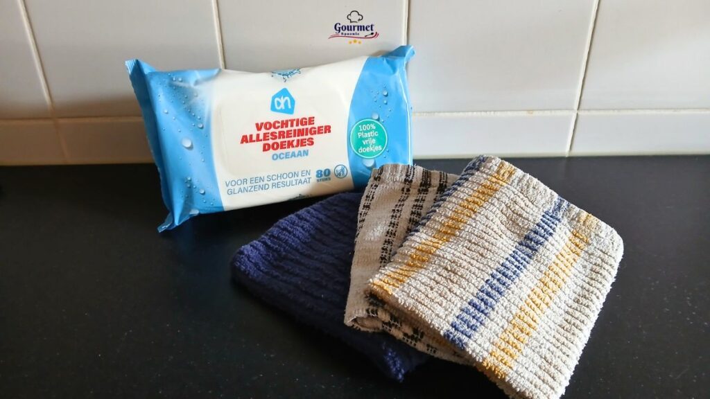 A pack of Ocean-scented cleaning wipes stands upright on a kitchen counter next to three folded, striped dishcloths in dark blue, beige, and white—offering practical cleaning cloths and wipes for easy kitchen hygiene tips against a white tiled backsplash.