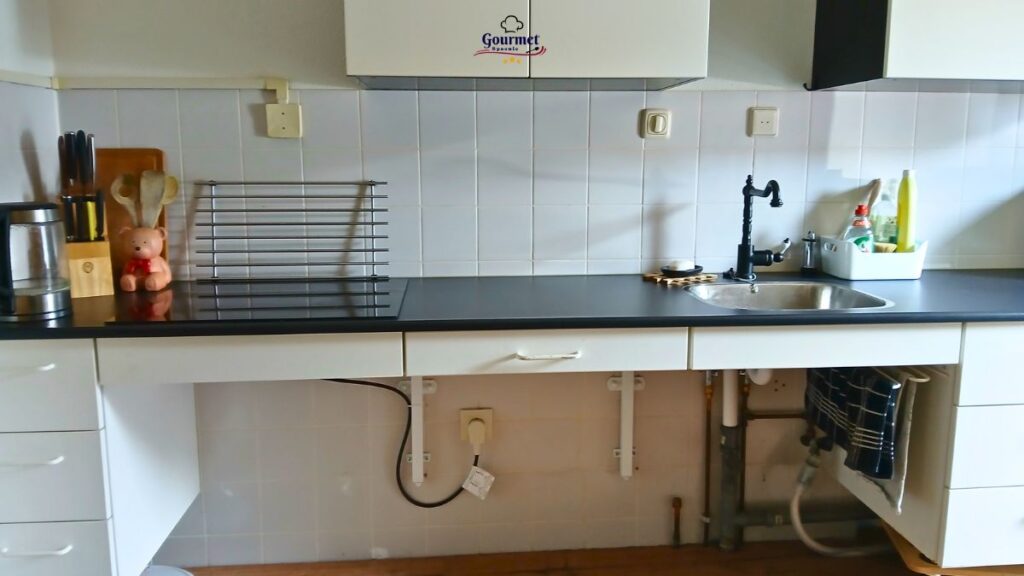 A clean kitchen countertop with minimal clutter features a knife block, utensils, a dish rack, a small teddy bear, a sink with a faucet, cleaning supplies, and exposed plumbing under the sink against a tiled backsplash—easy kitchen cleaning tips for spoonies.