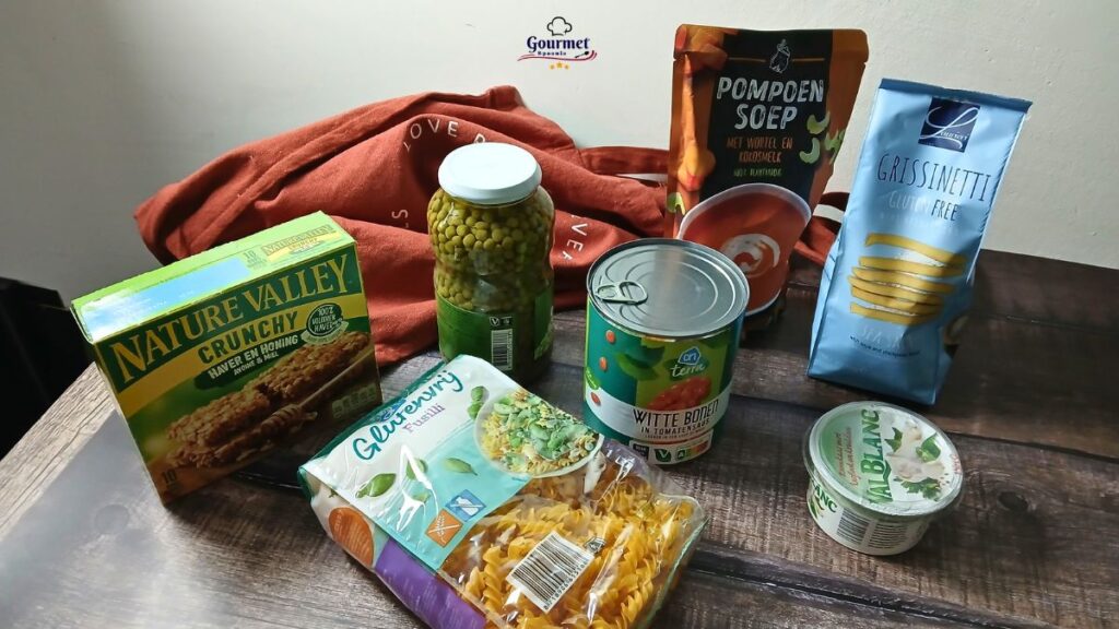 A selection of packaged foods on a wooden table, ideal as emergency meals for spoonies, including granola bars, pasta, soup, breadsticks, canned peas, preserved vegetables, and a tub of cream cheese. A red cloth shopping bag and Gourmet Spoonie logo appear in the background.