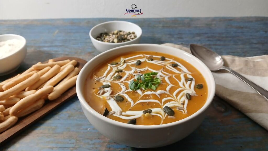 A bowl of creamy orange pumpkin soup, perfect for quick emergency meal ideas, is garnished with a white swirl and pumpkin seeds. It sits on a rustic blue table with breadsticks, a spoon, napkin, and a small bowl of seeds nearby.