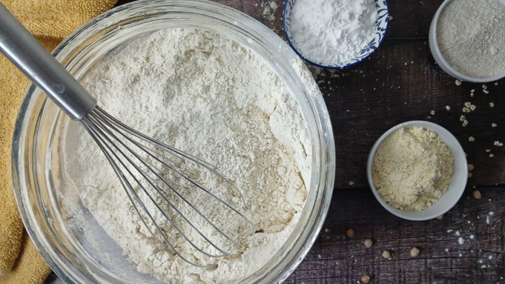 A metal whisk rests in a glass bowl of homemade gluten-free flour blend on a wooden surface, surrounded by small bowls containing flour and other dry baking ingredients. A yellow cloth is partially visible in the corner.