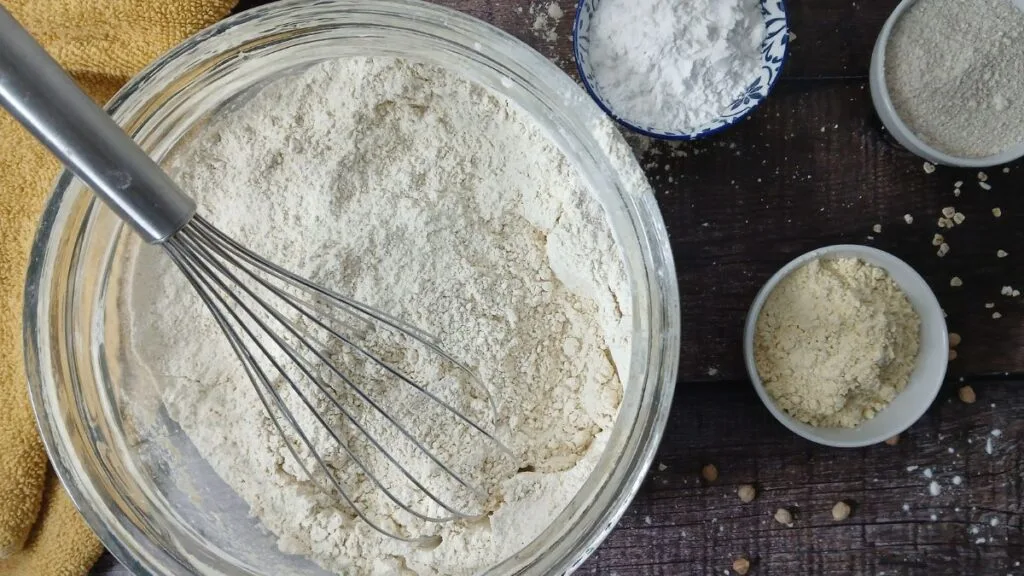 A metal whisk rests in a glass bowl of homemade gluten-free flour blend on a wooden surface, surrounded by small bowls containing flour and other dry baking ingredients. A yellow cloth is partially visible in the corner.
