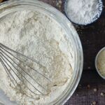 A metal whisk rests in a glass bowl of homemade gluten-free flour blend on a wooden surface, surrounded by small bowls containing flour and other dry baking ingredients. A yellow cloth is partially visible in the corner.