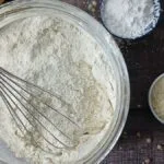 A metal whisk rests in a glass bowl of homemade gluten-free flour blend on a wooden surface, surrounded by small bowls containing flour and other dry baking ingredients. A yellow cloth is partially visible in the corner.