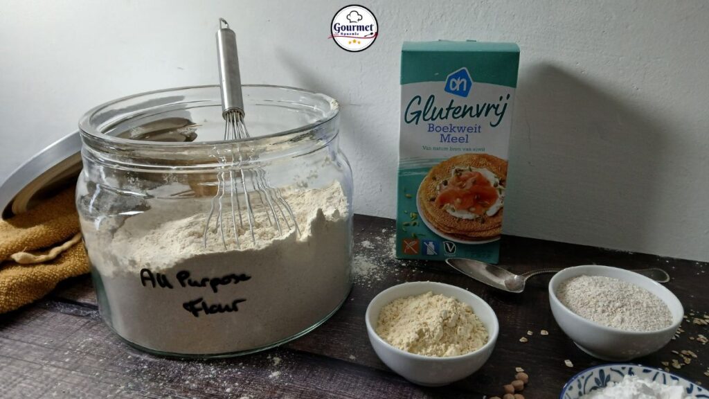 A jar labeled “All Purpose Flour” containing a homemade gluten-free flour blend with a whisk inside stands next to a box of gluten-free buckwheat flour. Small bowls of various gluten-free flours, are arranged on a dark wooden surface in front of a white wall.