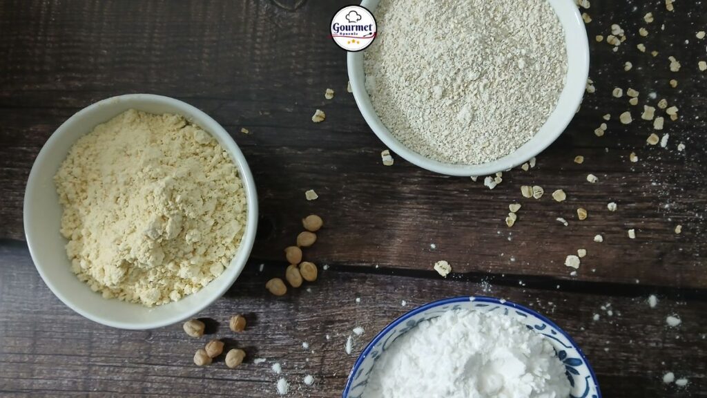 Three bowls with different flours—one with yellow chickpea flour and chickpeas, one with white tapioca starch, and one with oat flour—are arranged on a dark wooden surface, with some oats and chickpeas scattered around.