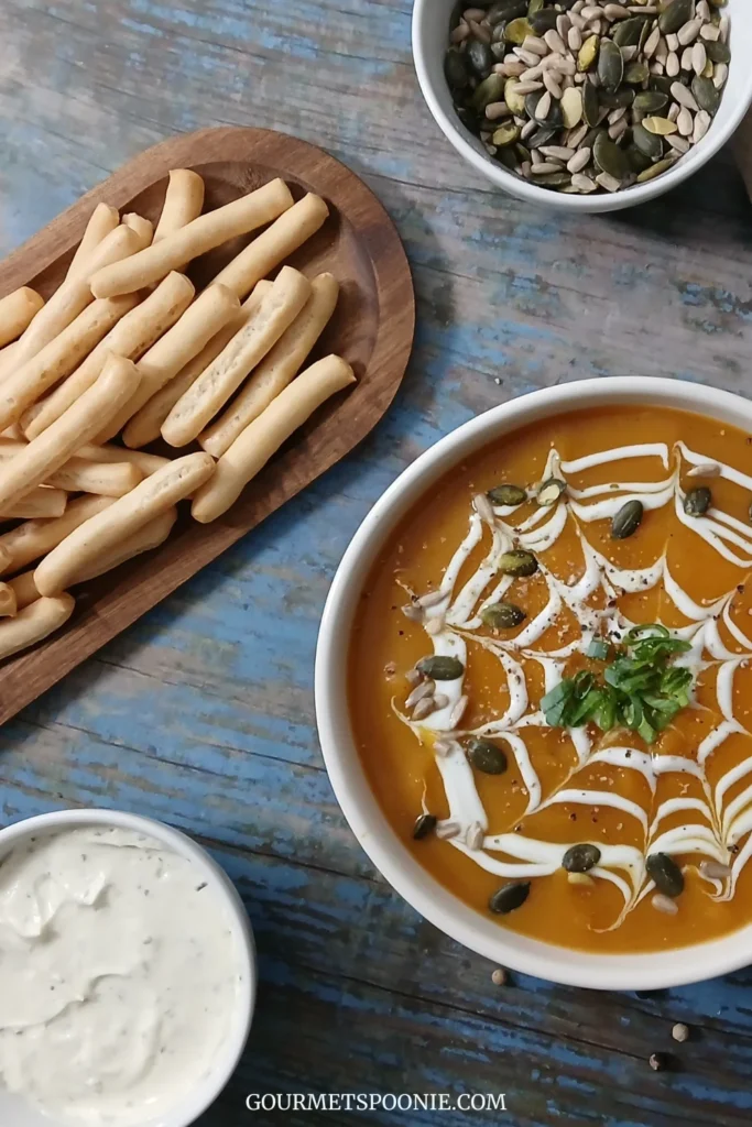 A bowl of soup with a creamy spiderweb design, a wooden dish with breadsticks, a bowl with seeds, and a creamy dip sit on a blue wooden table.
