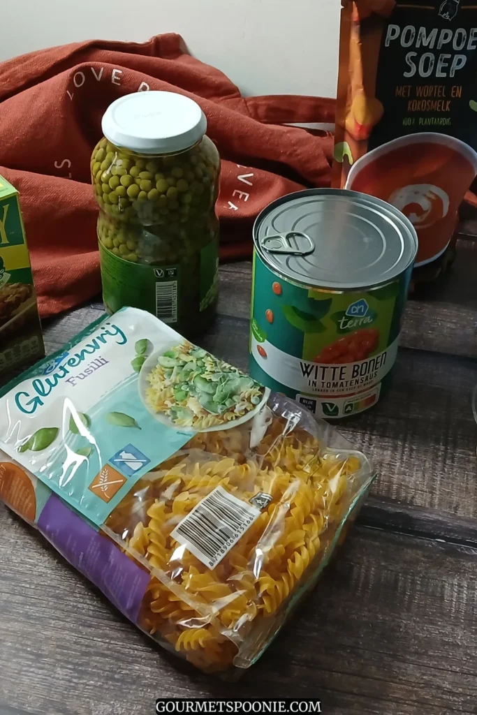 A selection of packaged foods on a wooden table, ideal as emergency meals for spoonies, including granola bars, pasta, soup, breadsticks, canned peas, preserved vegetables, and a tub of cream cheese. A red cloth shopping bag appears in the background.