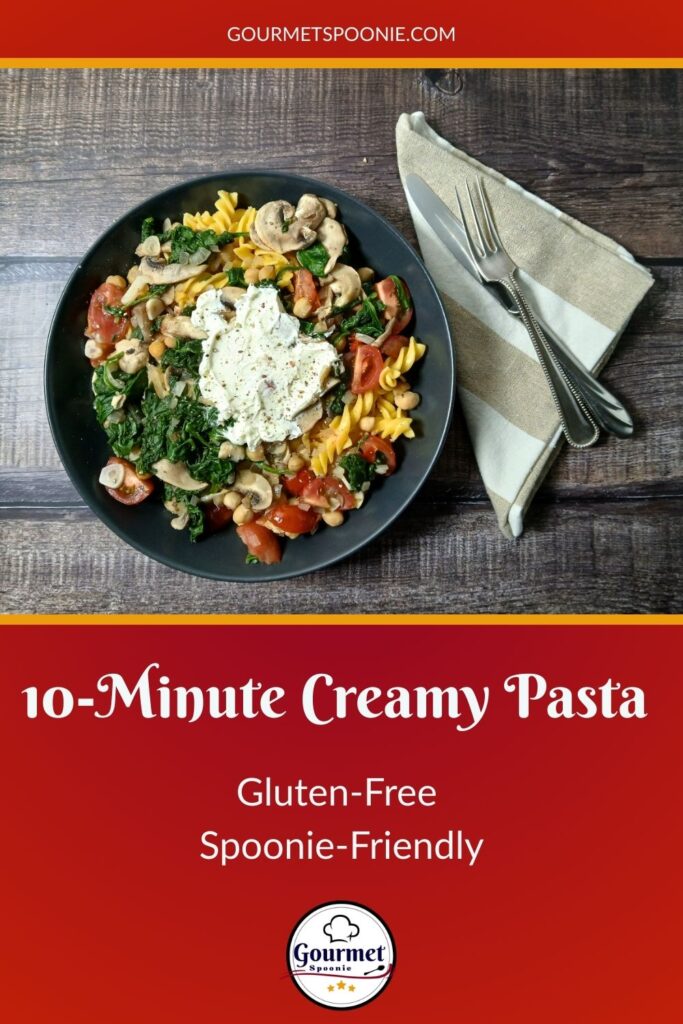 A black bowl filled with 10-Minute Creamy Pasta, mushrooms, spinach, and tomatoes sits on a wood surface next to a beige napkin with a fork and knife. Text reads: “10-Minute Creamy Pasta, Gluten-Free, Spoonie-Friendly—Gourmet Spoonie.”.