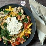 A black plate with Gluten-Free Creamy Pasta with Chickpeas & Spinach, topped with sautéed mushrooms, tomatoes, and a dollop of ricotta. A fork and knife rest on a folded beige napkin beside the plate on a wooden table.