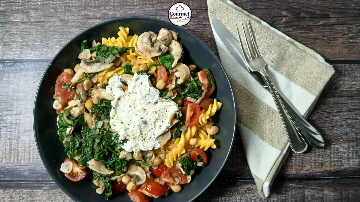A black plate with Gluten-Free Creamy Pasta with Chickpeas & Spinach, topped with sautéed mushrooms, tomatoes, and a dollop of ricotta. A fork and knife rest on a folded beige napkin beside the plate on a wooden table.