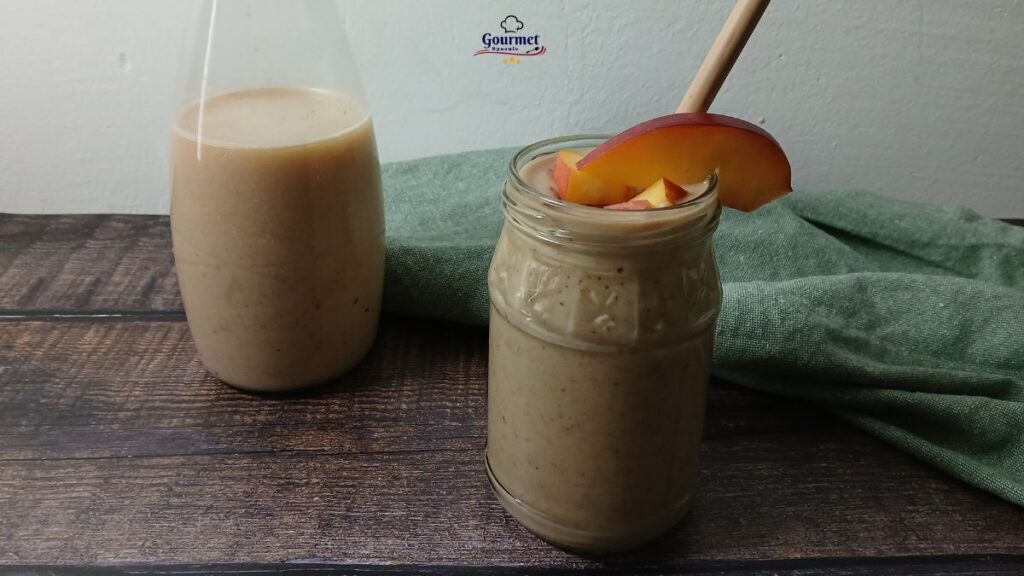A jar of healthy breakfast smoothie with oats and nuts, garnished with peach slices and a straw, sits next to a bottle of smoothie on a wooden table with a green cloth in the background.