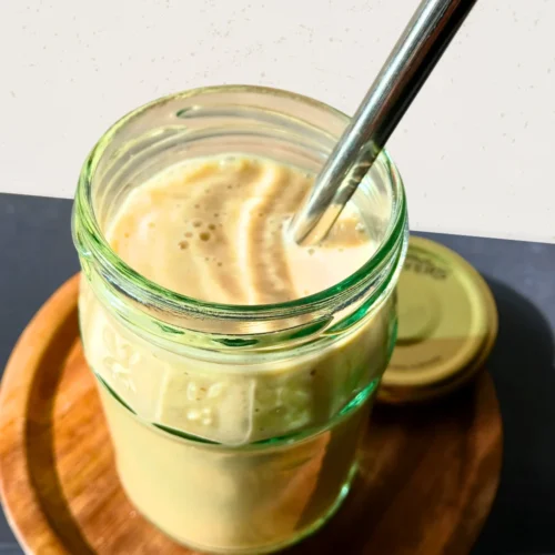 A glass jar filled with a breakfast smoothie with oats and nuts sits on a wooden coaster, with a metal straw and a jar lid resting nearby.