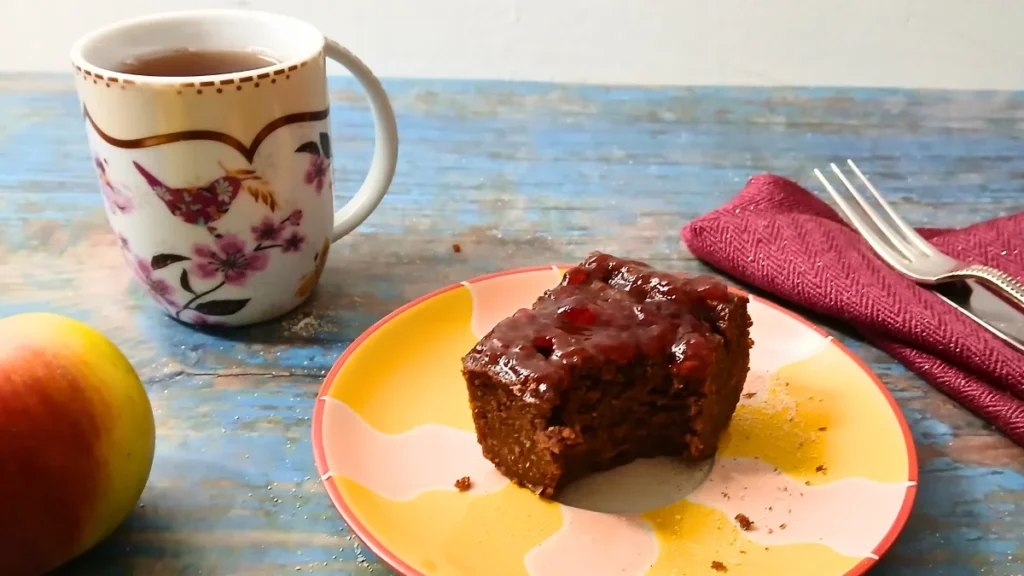 A cup of tea, a floral mug, and gluten-free chickpea brownies with jam on a colorful plate sit on a blue wooden table. An apple, red napkin, and silverware are nearby.