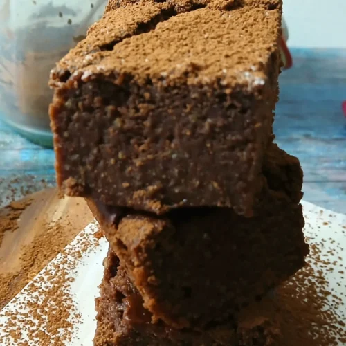 Three stacked gluten-free brownies dusted with cocoa powder sit on a white surface. In the background is a glass jar.