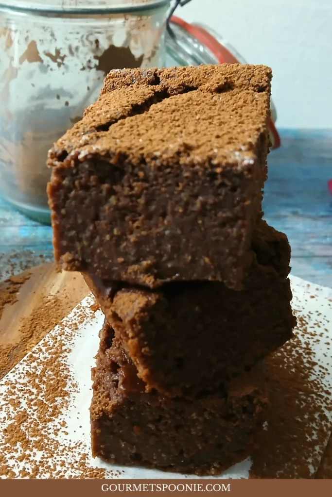 Three stacked gluten-free brownies dusted with cocoa powder sit on a white surface. In the background is a glass jar.