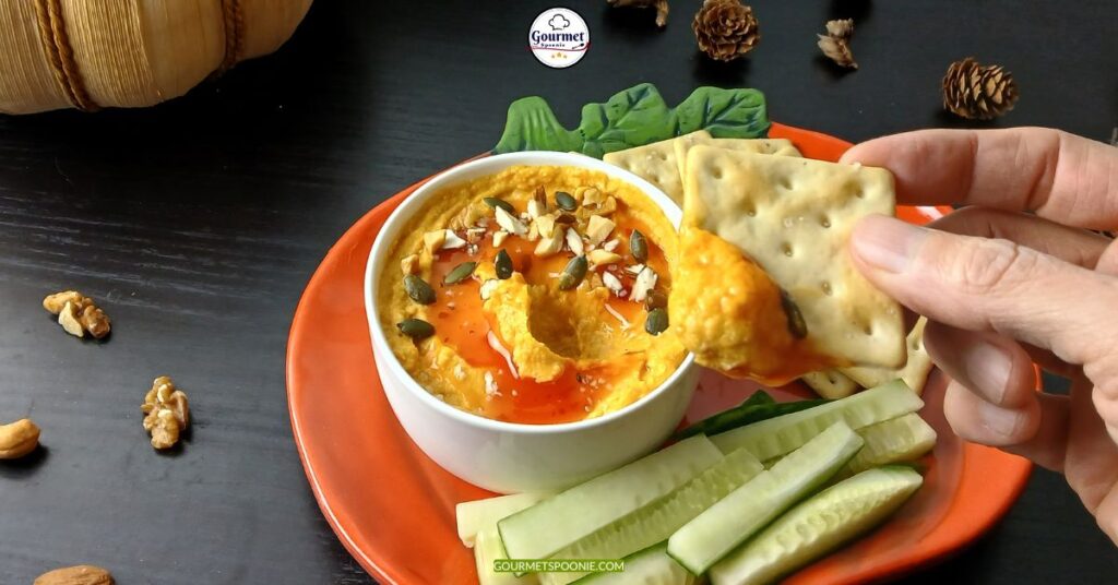 A hand dips a cracker into a bowl of creamy plant-based pumpkin dip, topped with seeds and chopped nuts, served with cucumber sticks on an orange plate. The background features nuts, pinecones, and a wooden basket.