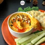 A hand dips a cracker into a bowl of creamy plant-based pumpkin dip, topped with seeds and chopped nuts, served with cucumber sticks on an orange plate. The background features nuts, pinecones, and a wooden basket.