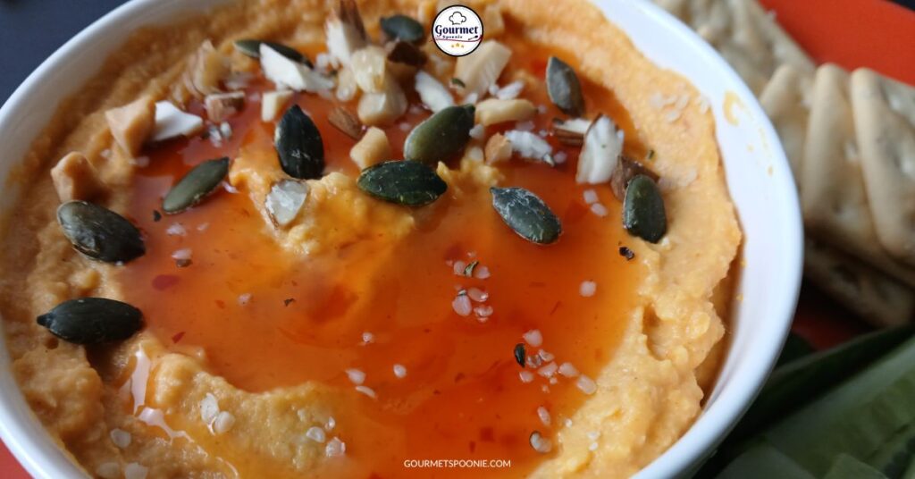 A close-up of a bowl of savoury pumpkin hummus topped with pumpkin seeds, chopped nuts, coarse salt, and a drizzle of sweet chili sauce. A few gluten-free crackers are visible on the side.