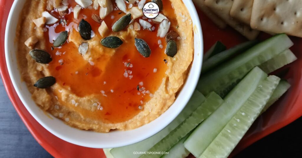 A bowl of butternut squash hummus topped with nuts, seeds, and sweet chili sauce is served on a red plate with sliced cucumbers and crackers on the side.